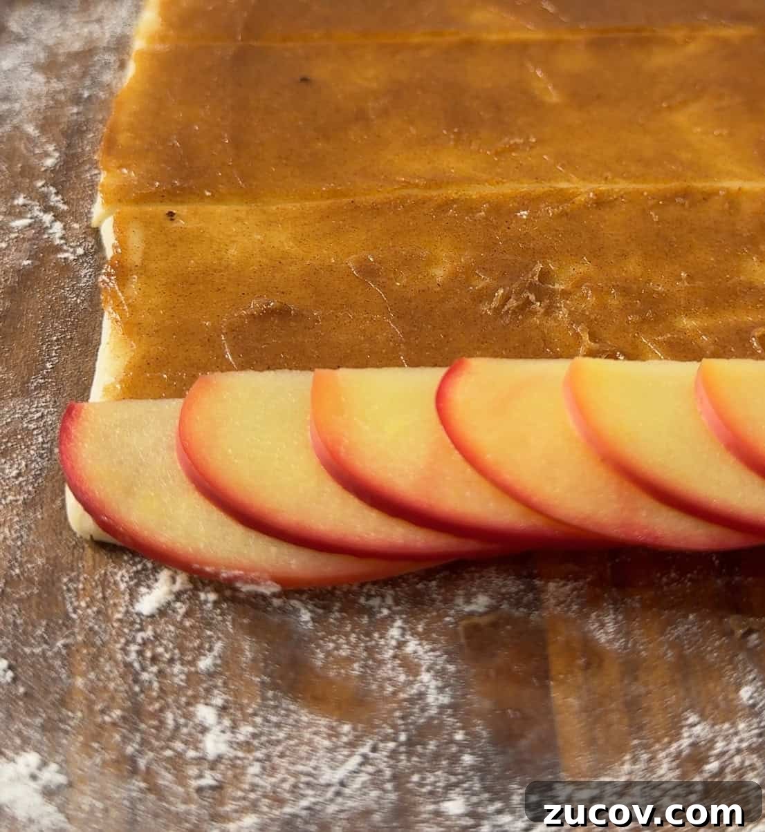 Apple Rose Pastry Recipe: Step-by-Step Homemade Apple Roses 3 Sliced red apples are neatly arranged on the edge of a rectangular piece of dough spread with brown filling, ready to be rolled on a floured surface.