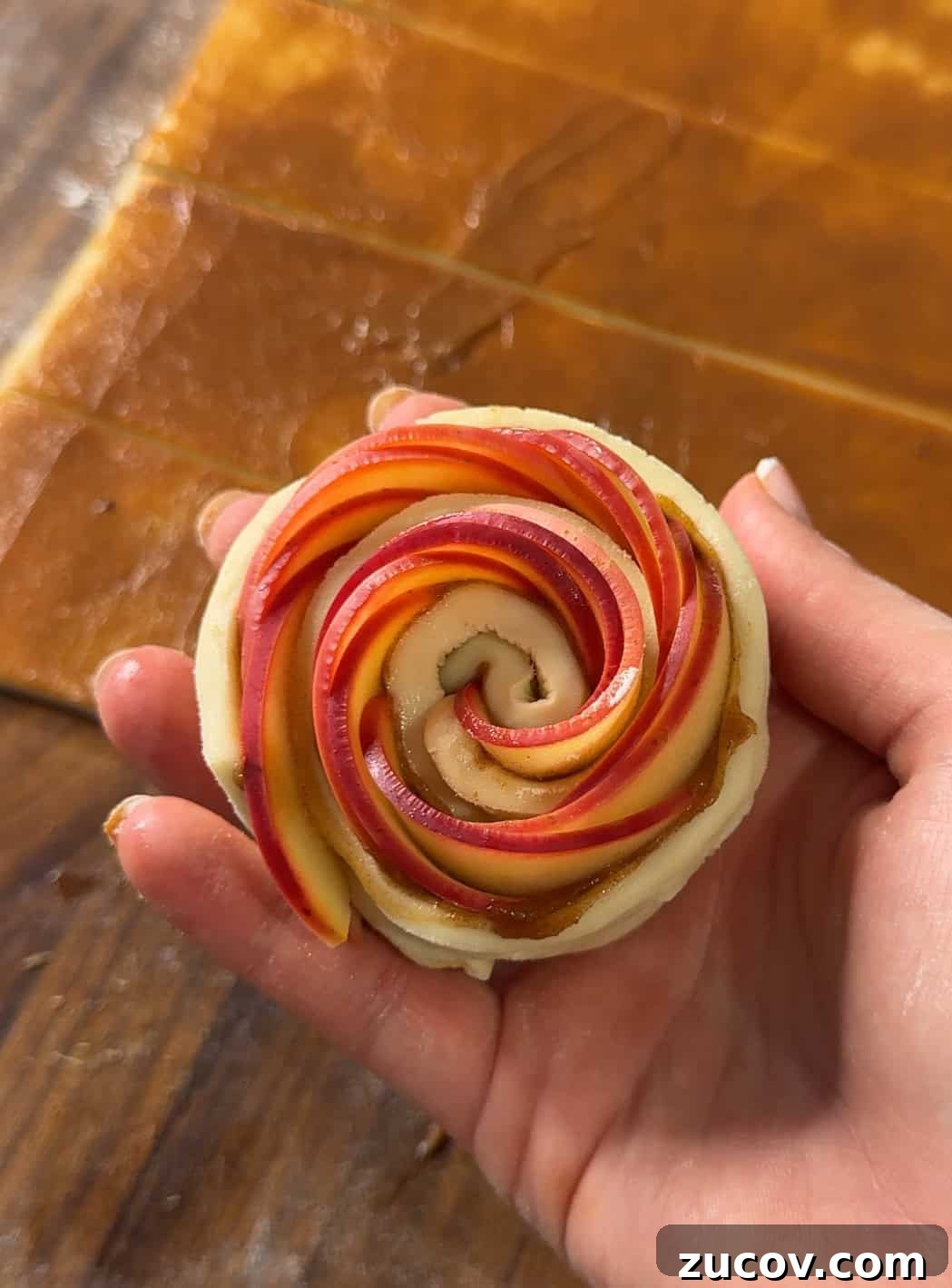 Apple Rose Pastry Recipe: Step-by-Step Homemade Apple Roses 4 A hand holds a pastry shaped like a rose, made from thin apple slices arranged in a spiral on top of dough. Strips of dough and apple filling are visible on the wooden surface in the background.