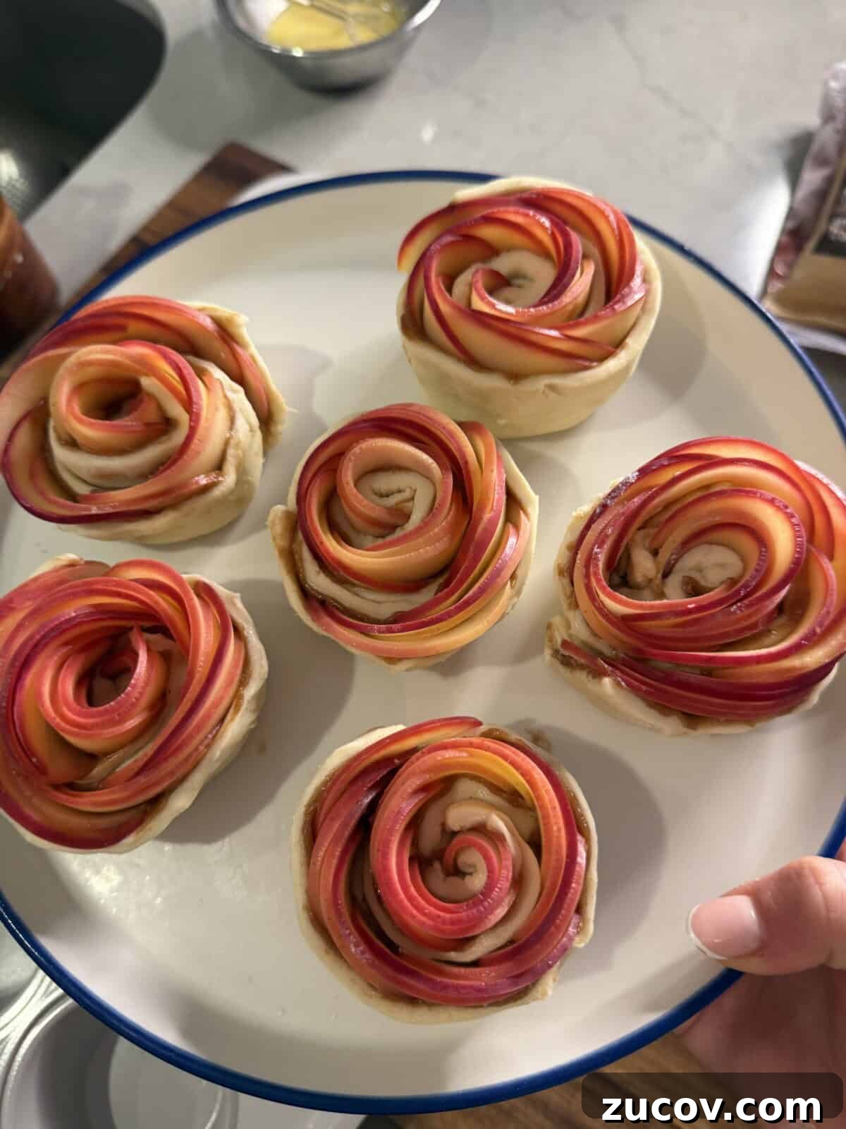Apple Rose Pastry Recipe: Step-by-Step Homemade Apple Roses 5 A hand holds a white plate with six pastries shaped like roses, made from thinly sliced red and pink apples arranged in spirals atop a light dough base. A kitchen counter with ingredients is visible in the background.