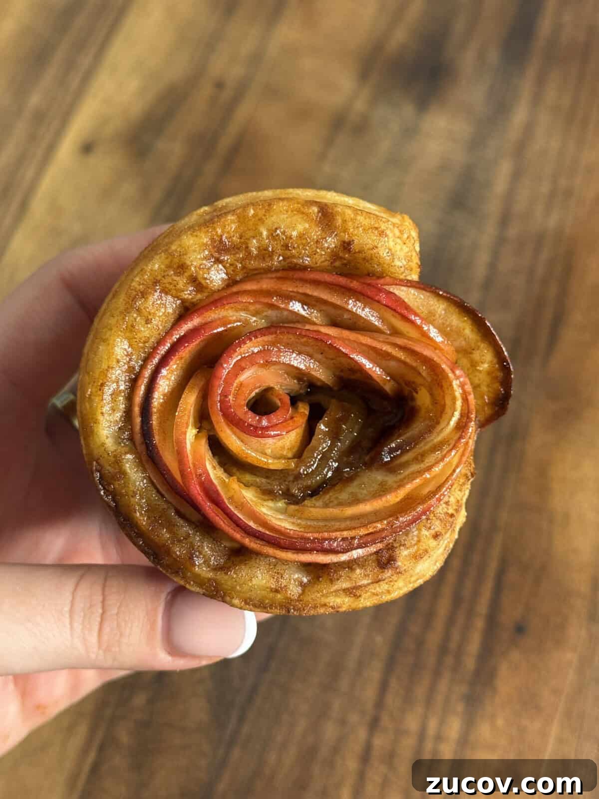 Apple Rose Pastry Recipe: Step-by-Step Homemade Apple Roses 6 A hand holds a pastry shaped like a rose, made from thinly sliced red apples arranged in spirals, with a golden-brown crust, against a wooden background.