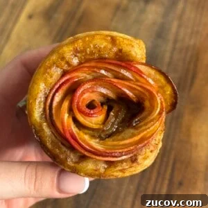 Apple Rose Pastry Recipe: Step-by-Step Homemade Apple Roses 7 A hand holds a pastry shaped like a rose, made from thinly sliced red apples arranged in spirals, with a golden-brown crust, against a wooden background.
