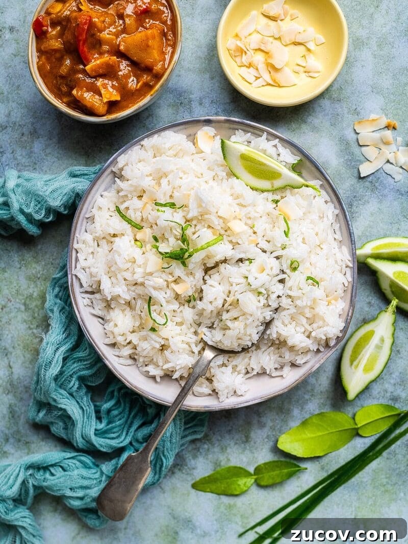 Coconut Rice Recipe: Creamy, Fragrant Side Dish in 30 Minutes 3 A bowl of coconut rice garnished with lime wedges and sliced almonds, with a spoon resting inside. Nearby are a small bowl of curry, a dish of almonds, lime slices, and herbs on a textured surface.