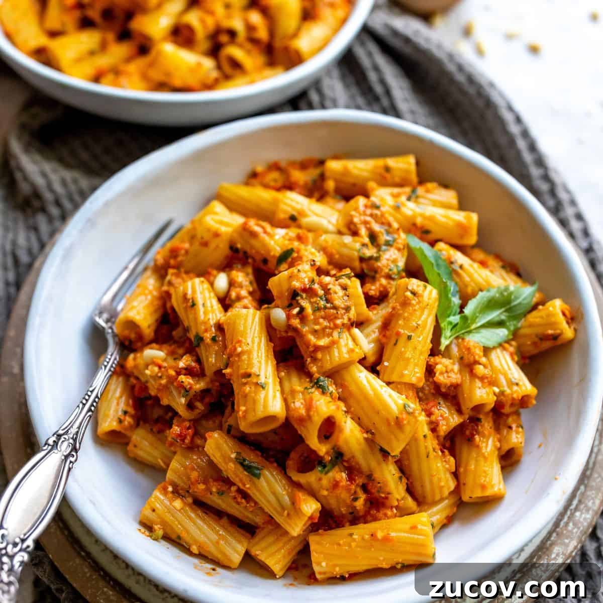 Roasted Red Pepper Pesto Recipe for Pasta, Sandwiches & Dips 2 White bowl with pesto and red peppers and noodles. A black fork in the bowl with grey towel.