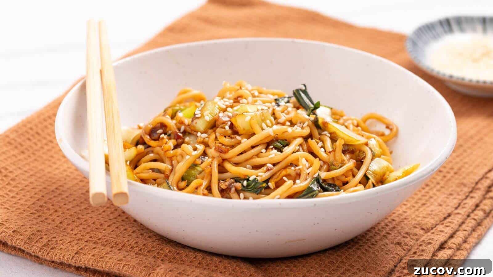 17 Comforting Side Dishes That Steal the Show 4 A bowl of stir-fried noodles with vegetables, topped with sesame seeds, placed on a brown cloth napkin. Chopsticks rest on the bowl's edge, and a small dish is visible in the background.