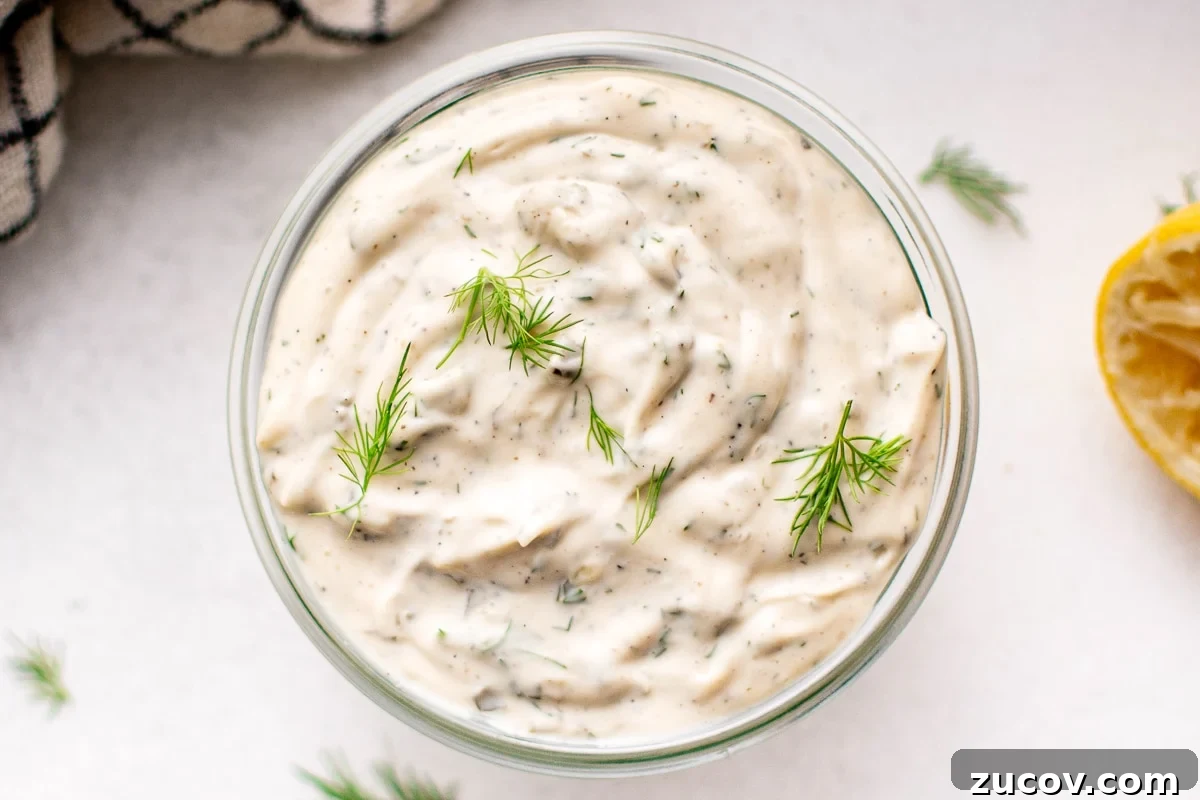 Creamy Homemade Tartar Sauce Recipe for Fish and Seafood 2 Overhead view of a glass jar filled with tartar sauce, garnished with dill sprigs, and surrounded by fresh herbs and a squeezed lemon half.