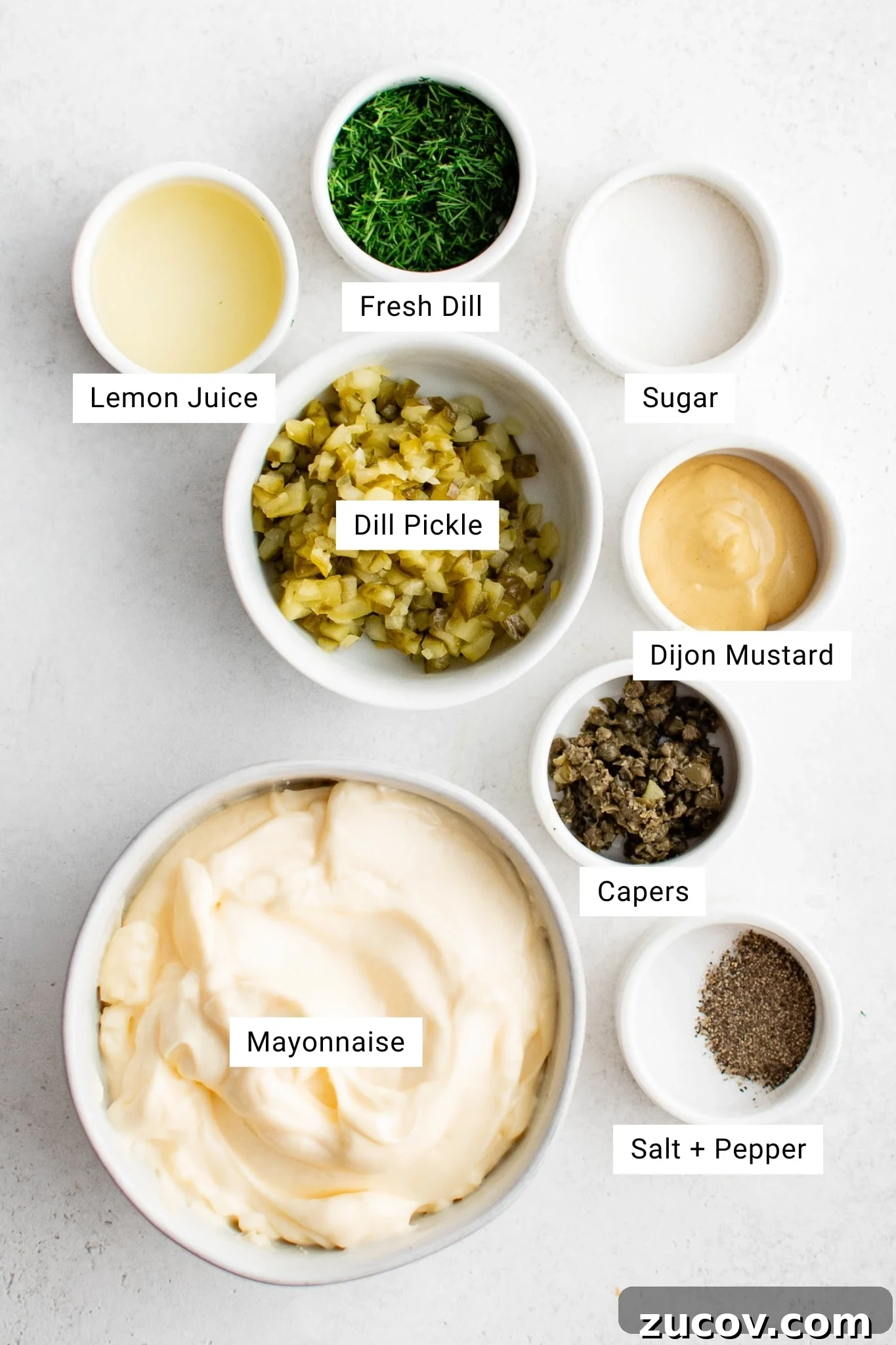Creamy Homemade Tartar Sauce Recipe for Fish and Seafood 3 Overhead view of tartar sauce ingredients in white bowls: mayonnaise, dill pickle, fresh dill, lemon juice, Dijon mustard, sugar, capers, salt and pepper.
