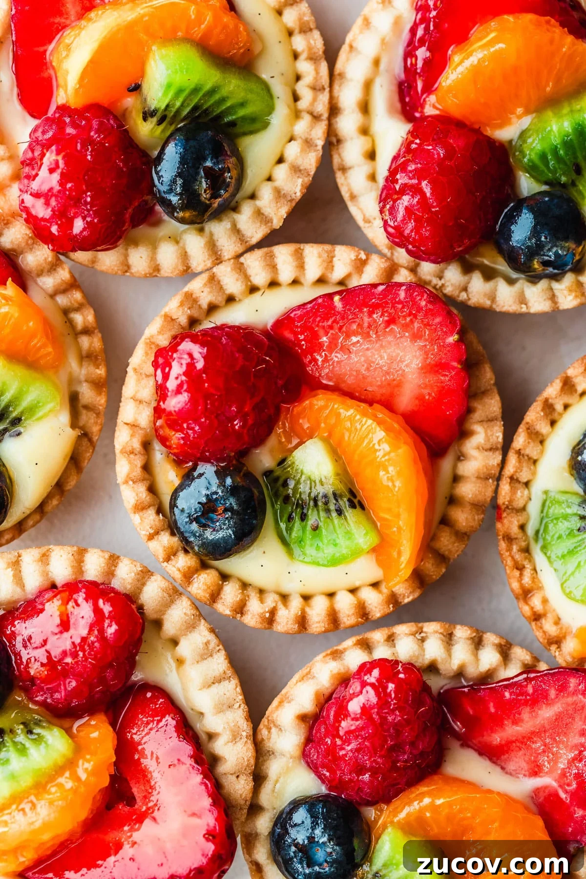 Fresh Fruit Tartlets with Silky Pastry Cream Recipe 2 Rows of mini fruit tartlets with berries, kiwi, and orange slices on a white counter.