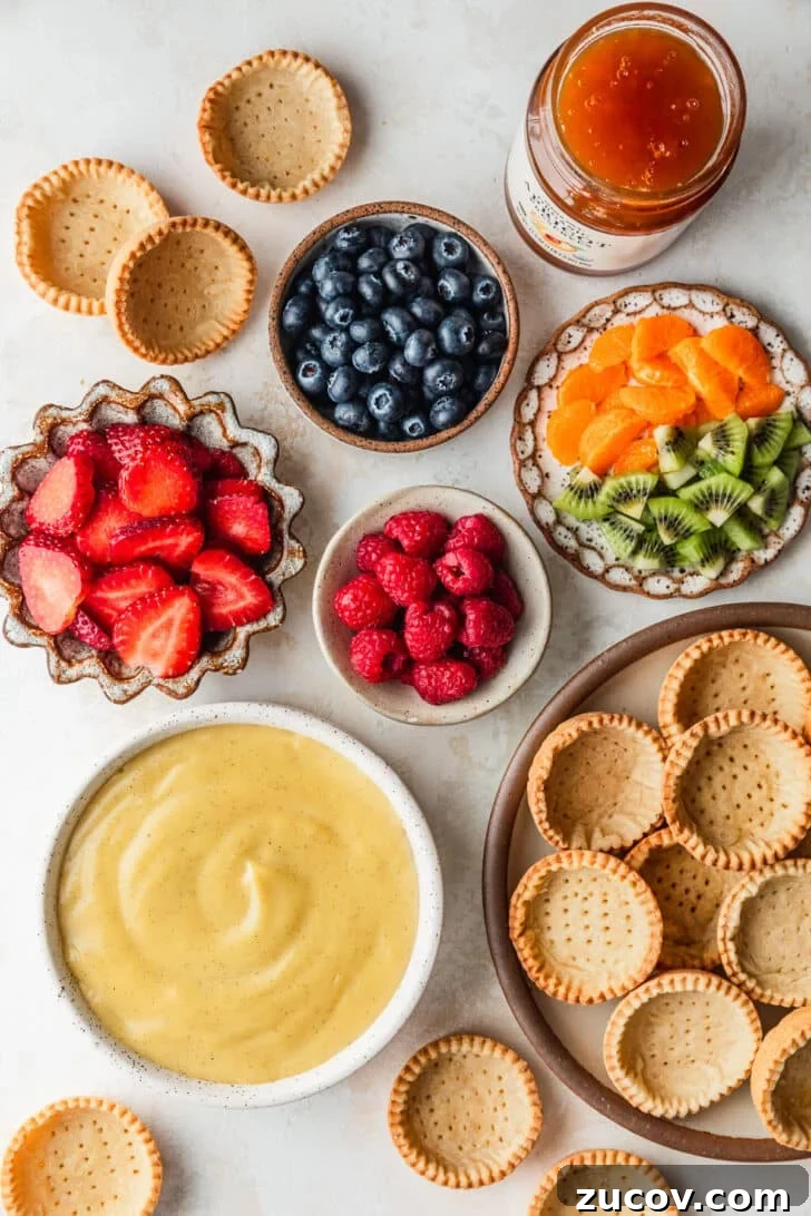 Fresh Fruit Tartlets with Silky Pastry Cream Recipe 4 White and brown bowls of ingredients for fruit tartlets like tart crusts, pastry cream, berries, kiwi, and oranges next to a jar of apricot jam with a tan backdrop.