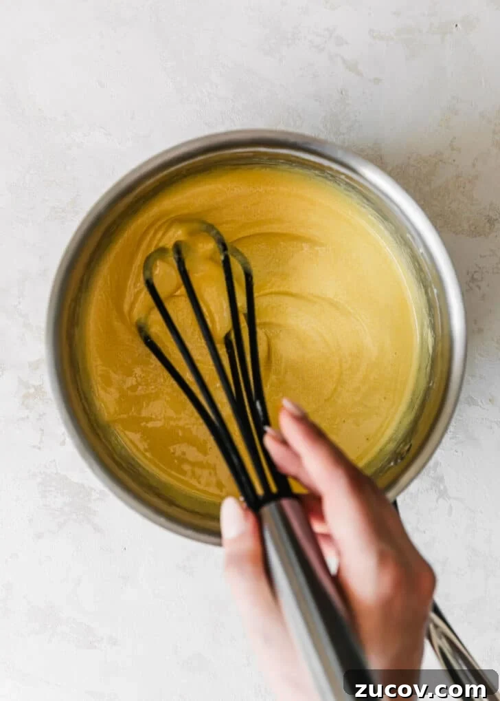 Fresh Fruit Tartlets with Silky Pastry Cream Recipe 7 A hand using a whisk to beat egg yolks, sugar, and cornstarch in a silver pot.