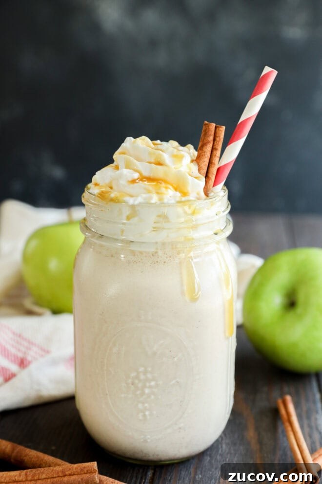 A mason jar filled with a creamy milkshake, topped with whipped cream, caramel drizzle, two cinnamon sticks, and a red-striped straw.