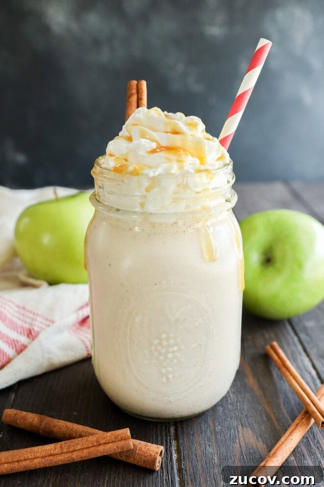 A mason jar filled with a creamy apple milkshake topped with whipped cream, caramel drizzle, and cinnamon sticks, with a red-and-white striped straw.
