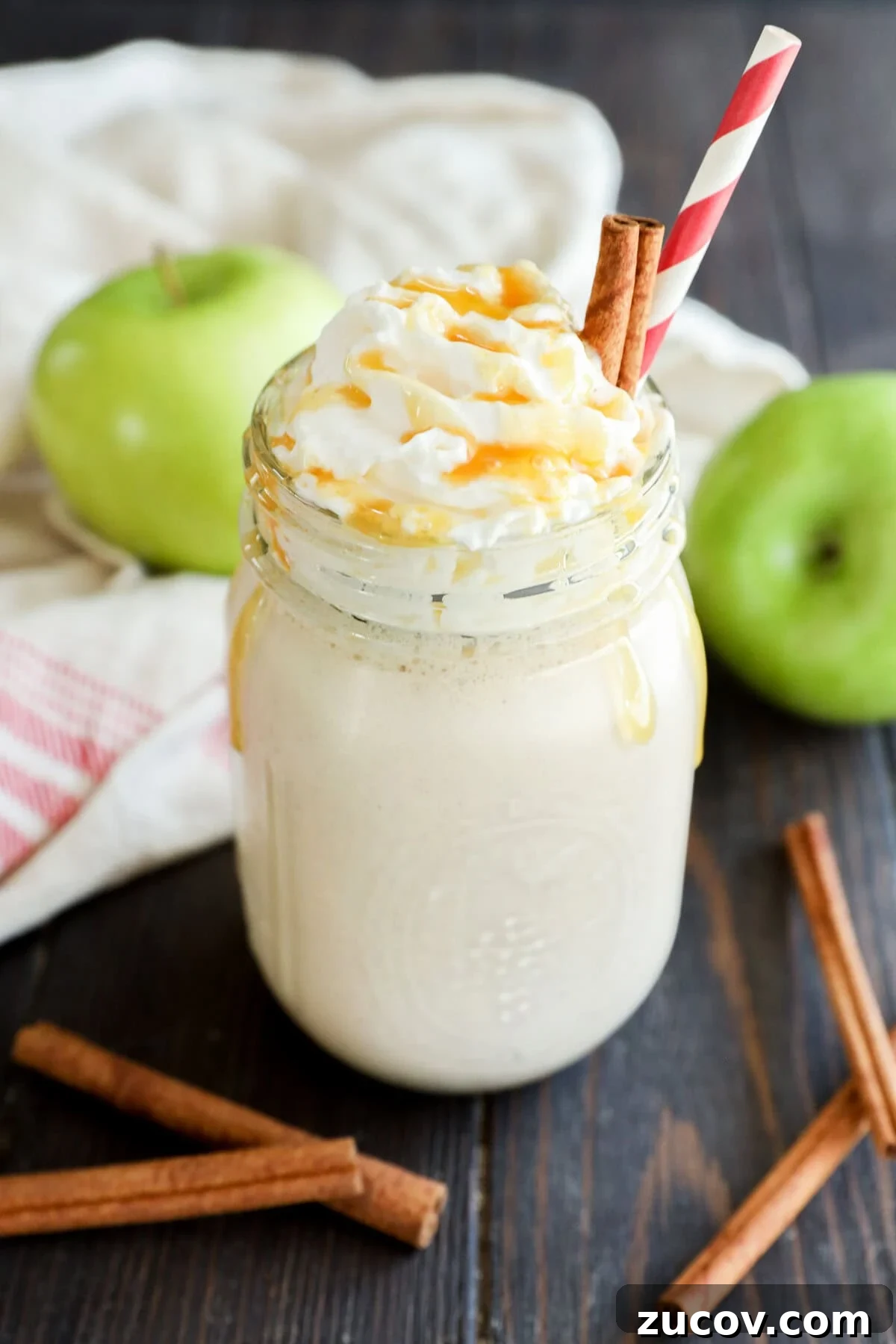 A creamy smoothie topped with whipped cream and caramel sauce in a mason jar, garnished with cinnamon sticks and a red-striped straw.