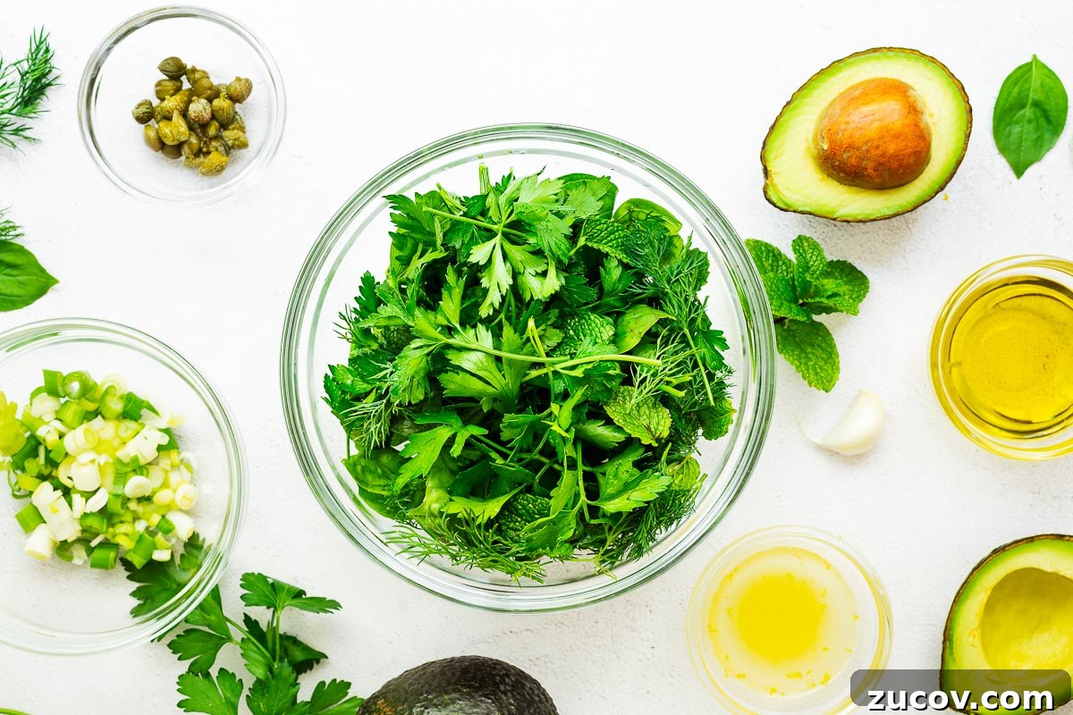 Creamy Avocado Green Goddess Dressing Recipe for Salads and Bowls 4 Chopped green herbs surrounded by the rest of the ingredients for vegan green goddess dressing, including sliced avocado, olive oil, lemon juice and zest, chopped green onions and capers.