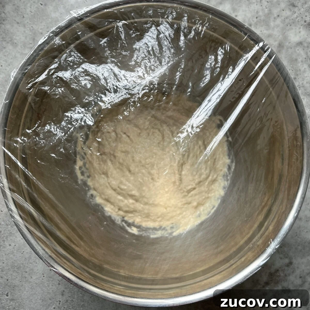 No-Knead Focaccia Recipe: Crispy, Olive Oil-Rich Bread in One Pan 11 A metal bowl containing Easy No-Knead Focaccia dough, covered with plastic wrap, rests on a gray countertop.
