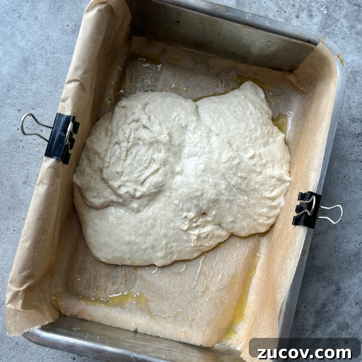 No-Knead Focaccia Recipe: Crispy, Olive Oil-Rich Bread in One Pan 12 Focaccia dough is transferred to a rectangular baking pan, with parchment paper secured with binder clips. The dough sits atop a layer of olive oil.