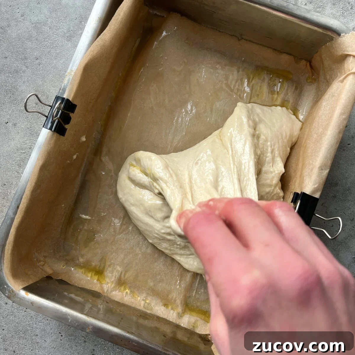 No-Knead Focaccia Recipe: Crispy, Olive Oil-Rich Bread in One Pan 13 A hand stretches and folds sticky Easy No-Knead Focaccia dough inside a parchment-lined metal baking tray secured with binder clips.