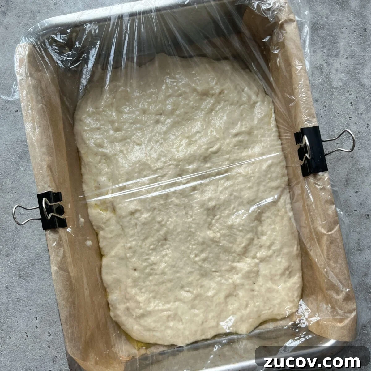 No-Knead Focaccia Recipe: Crispy, Olive Oil-Rich Bread in One Pan 14 A rectangular baking pan lined with parchment paper holds Easy No-Knead Focaccia dough covered with plastic wrap to rise, secured at the sides with binder clips.