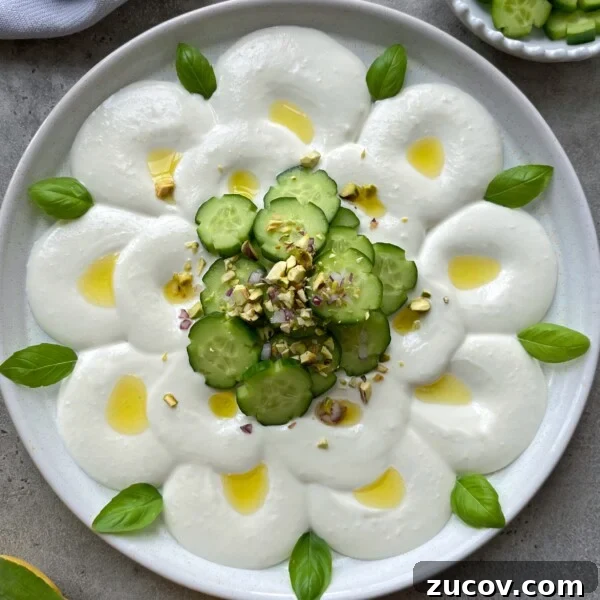 No-Knead Focaccia Recipe: Crispy, Olive Oil-Rich Bread in One Pan 17 A creamy white feta cheese dip is arranged in a flower pattern, topped with cucumber slices in the center, drizzled with olive oil, chopped pistachios, red onion, and garnished with fresh basil leaves.