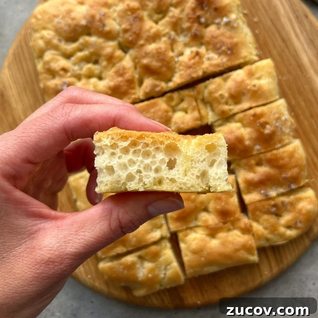 No-Knead Focaccia Recipe: Crispy, Olive Oil-Rich Bread in One Pan 3 A hand holds a slice of airy, golden focaccia bread above a wooden board with more focaccia pieces. This delicious bite showcases the bread’s light, fluffy texture—perfect for adding to your favorite food recipes or sharing at casual gatherings.