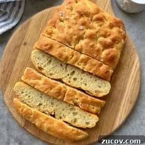 No-Knead Focaccia Recipe: Crispy, Olive Oil-Rich Bread in One Pan 21 A sliced loaf of focaccia bread with a golden crust, displayed on a round wooden cutting board.
