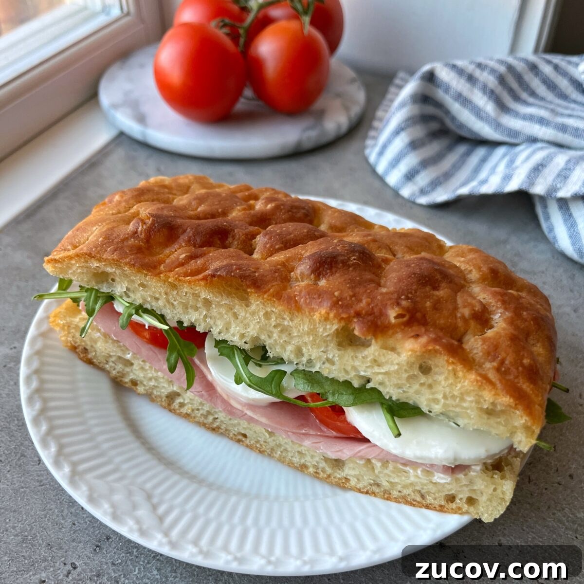 No-Knead Focaccia Recipe: Crispy, Olive Oil-Rich Bread in One Pan 5 A sandwich with ham, mozzarella, tomato, and arugula on Easy No-Knead Focaccia sits on a white plate.