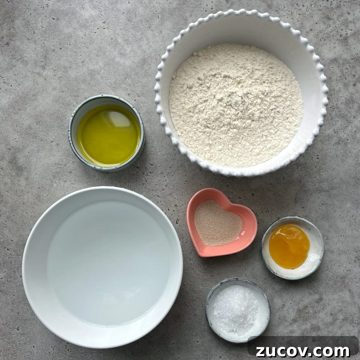 No-Knead Focaccia Recipe: Crispy, Olive Oil-Rich Bread in One Pan 6 A bowl of flour, olive oil, water, yeast in a heart-shaped dish, honey, and sea salt, arranged on a gray surface.