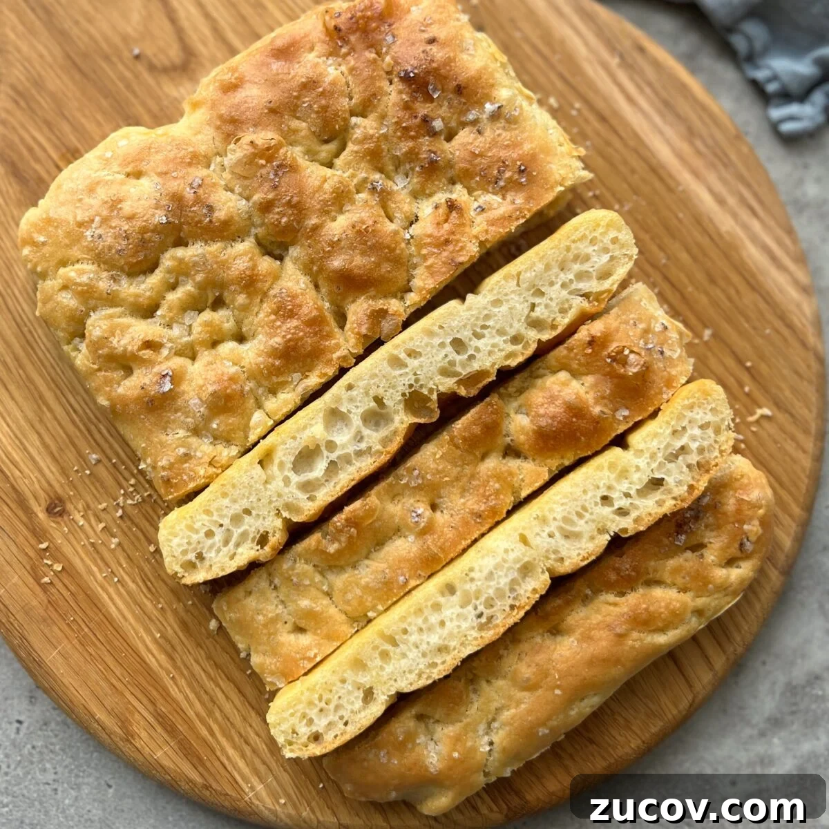 No-Knead Focaccia Recipe: Crispy, Olive Oil-Rich Bread in One Pan 7 A rectangular loaf of focaccia bread, featured in many food recipes, sits sliced on a round wooden board. The bread has a golden crust and a coarse, airy crumb.