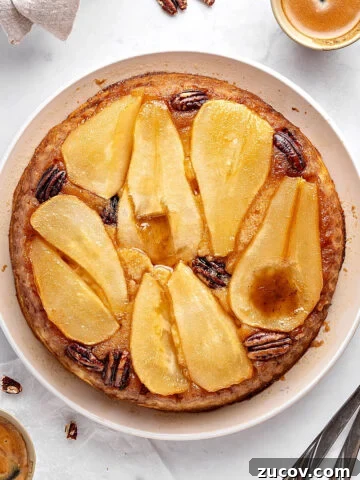 Top view of pear upside down cake on a white plate. Pecans, coffee cups, silver forks. Whitish background.