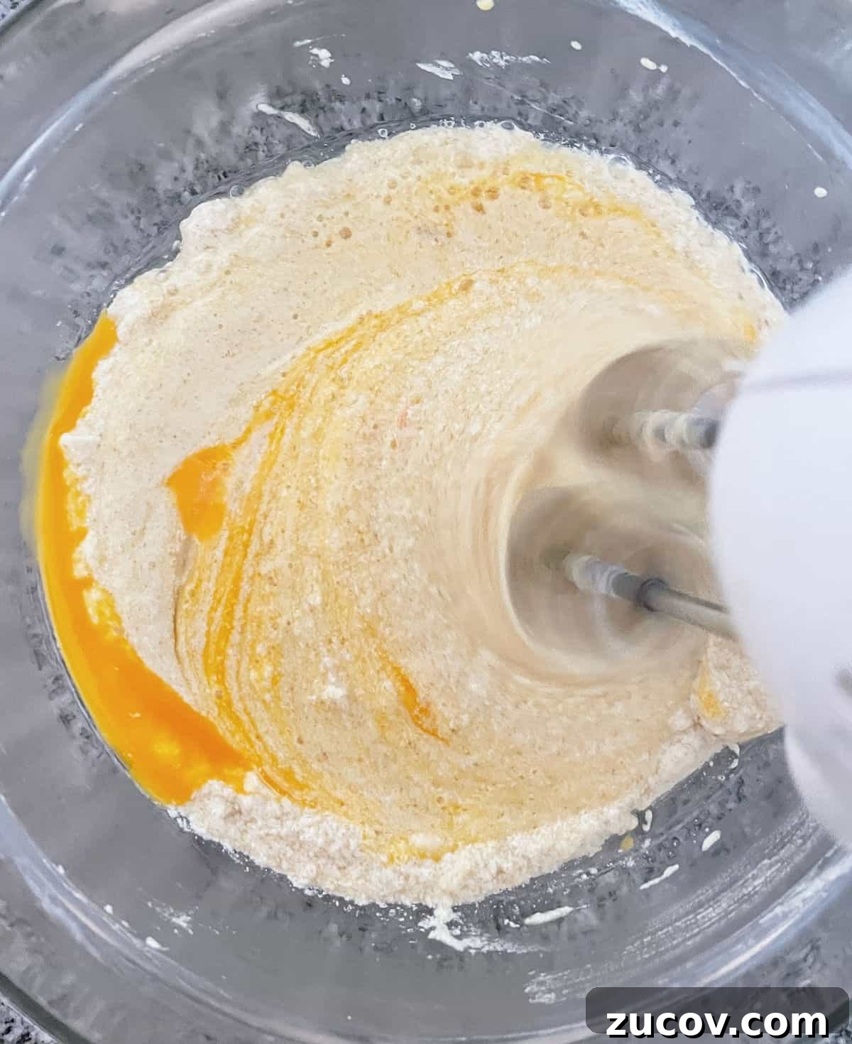 Moist Pear Cake Recipe with Cinnamon Glaze 5 Beating egg into cake batter in a glass bowl on a grey surface.