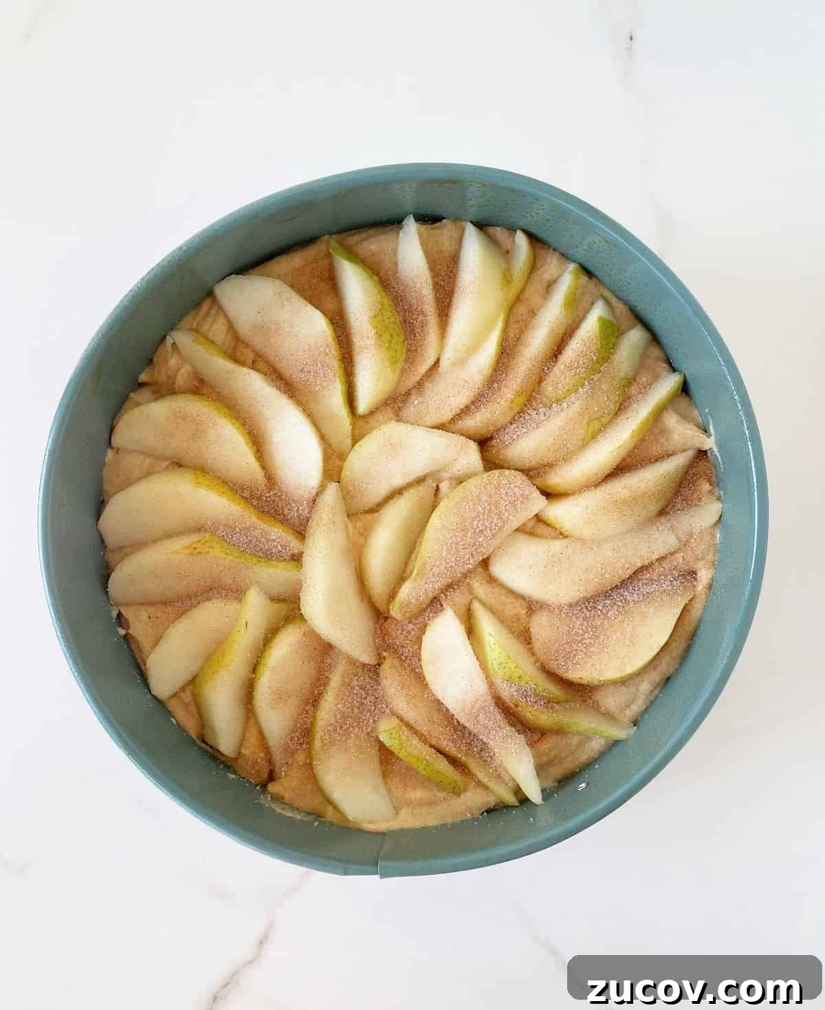 Moist Pear Cake Recipe with Cinnamon Glaze 8 Pear slices with cinnamon sugar on cake batter on a teal round pan on white marble surface.