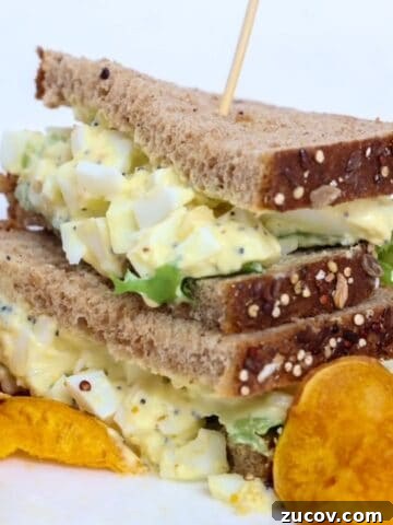 Two halves of an egg salad sandwich stacked, on a plate with sweet potato chips.