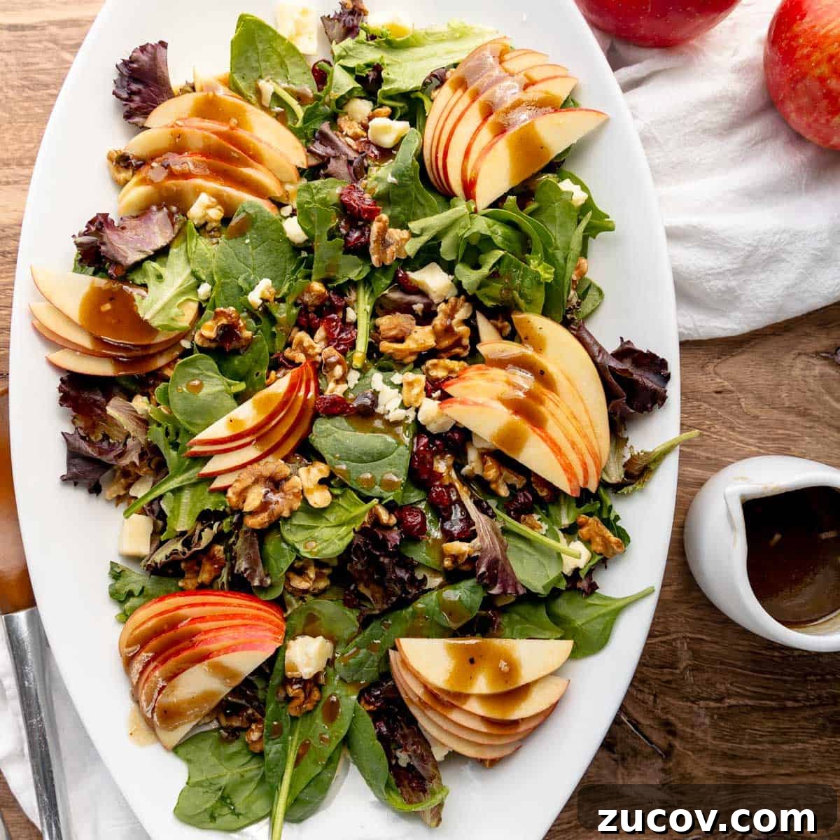 Crisp Apple and Walnut Salad with Maple Dijon Dressing 2 A white oval plate with a mixed greens salad topped with apple slices, walnuts, cranberries, and bits of cheese. A small white pitcher with dressing is on the side. Two red apples and a napkin are in the background.