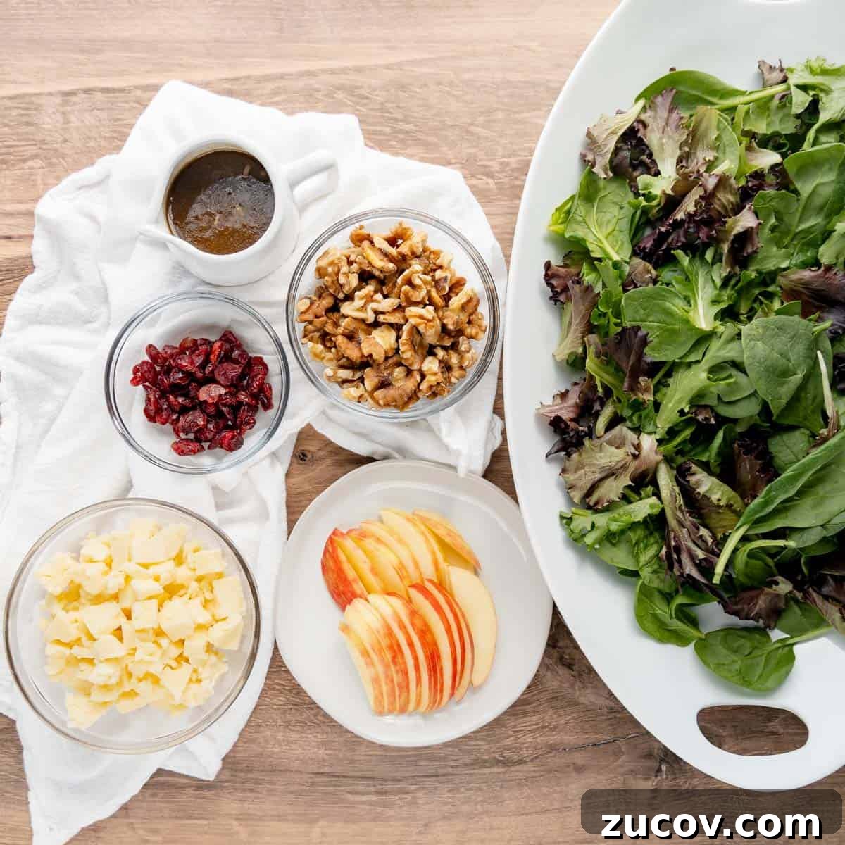 Crisp Apple and Walnut Salad with Maple Dijon Dressing 3 A large bowl of mixed greens with bowls of dressing, walnuts, dried cranberries, cheese, and apple slices arranged on a wooden surface.