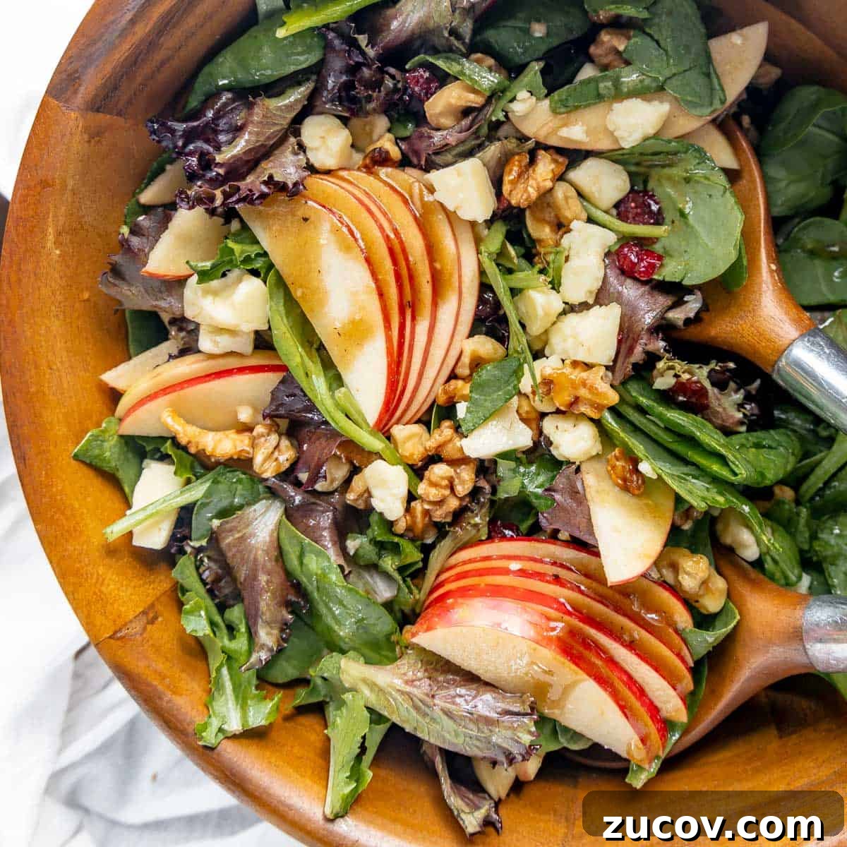 Crisp Apple and Walnut Salad with Maple Dijon Dressing 7 A wooden bowl filled with a mixed green salad featuring apple slices, walnuts, cheese cubes, and a light dressing, with wooden salad servers.