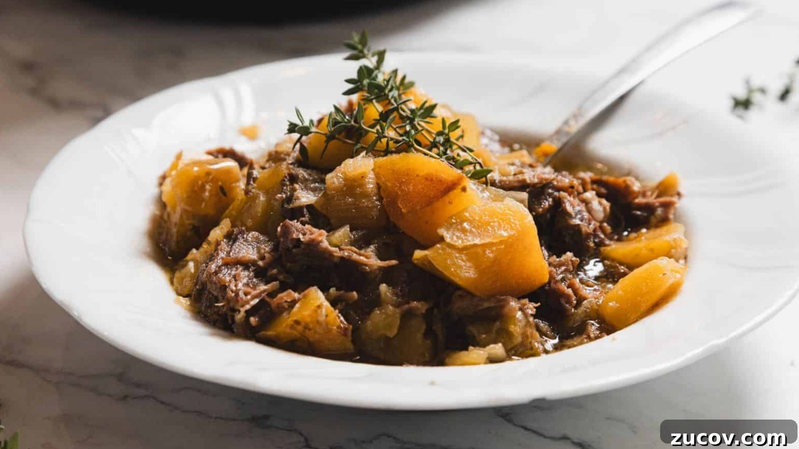 15 Irresistible St. Patrick’s Day Beef Recipes to Make Again and Again 3 A white plate containing a stew made of tender shredded meat and chunks of potatoes, garnished with sprigs of fresh thyme. A spoon rests on the side of the dish, placed on a marble surface.