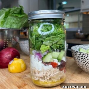A mason jar filled with layered salad ingredients, including shredded chicken, dressing, cherry tomatoes, olives, cucumbers, red onions, and chopped lettuce. The jar sits on a wooden counter with vegetables in the background.