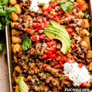 baking sheet with tater tots topped with ground beef, melted cheese, diced tomatoes, sour cream, and avocado slices