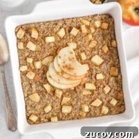 Baked apple oatmeal topped with apple slices in a square white baking dish with apples and syrup on the side.