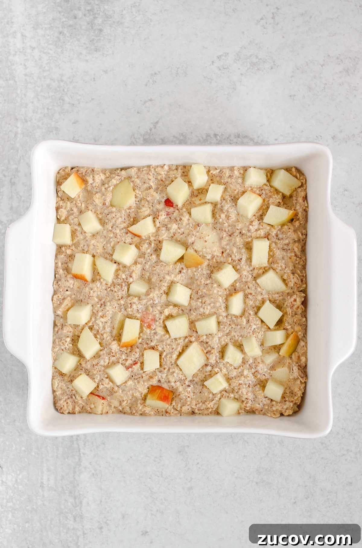 Oatmeal batter in a square white baking dish sprinkled with apple pieces.