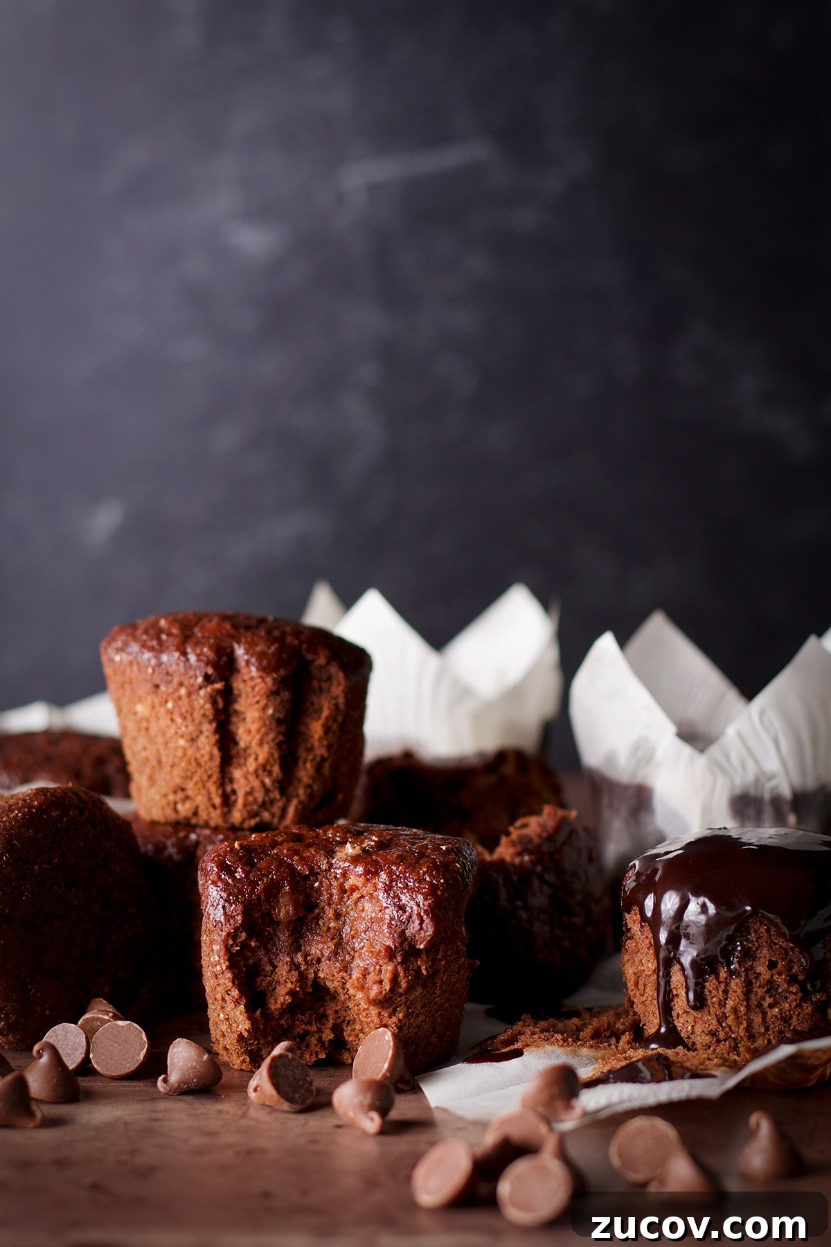 Chocolate Banana Muffins with Oats and Dark Cocoa 2 Several iced chocolate banana breakfast muffins on a wood table with chocolate chips scattered all around them.
