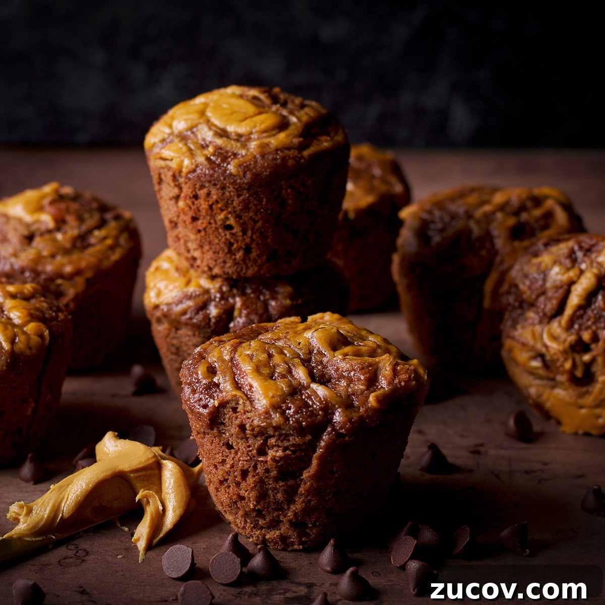 Chocolate Banana Muffins with Oats and Dark Cocoa 3 Chocolate banana muffins with a peanut butter swirl stacked on a table with a peanut butter covered knife laying next to them.